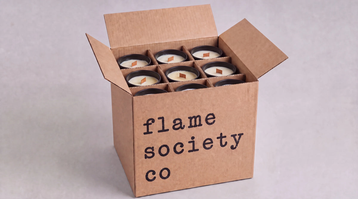 An open brown cardboard box printed with the text 'flame society co' holds six small candles arranged in a grid. The candles have white wax and dark lids, photographed on a light gray background.