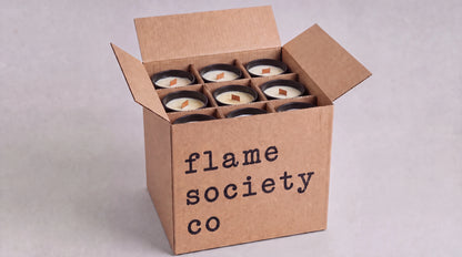 An open brown cardboard box printed with the text 'flame society co' holds six small candles arranged in a grid. The candles have white wax and dark lids, photographed on a light gray background.