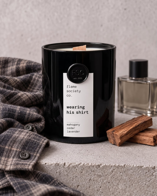 Wearing His Shirt candle in a glossy black glass jar with wooden wick, styled with a plaid flannel shirt, cologne bottle and cedar wood on concrete — mahogany, cedar and lavender — Flame Society Co.