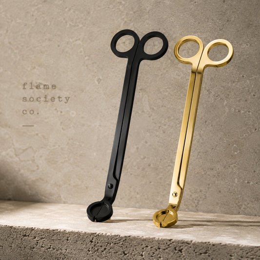 Flame Society Co. wick trimmers in matte black and polished gold leaning against travertine stone