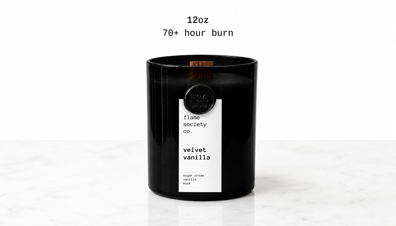 A black glass jar candle with a white label reading 'flame society co. velvet vanilla' and a small wooden wick, shown on a white marble surface against a white background with text above reading '12oz 70+ hour burn'.