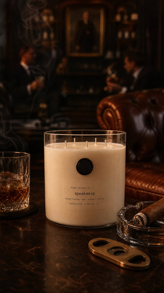 A large white candle in a clear glass jar sits on a dark wooden bar table, surrounded by a glass of whiskey, a cigar in a glass holder, and a brass cigar cutter. The background is a dimly lit, moody bar interior with leather seating and blurred figures.
