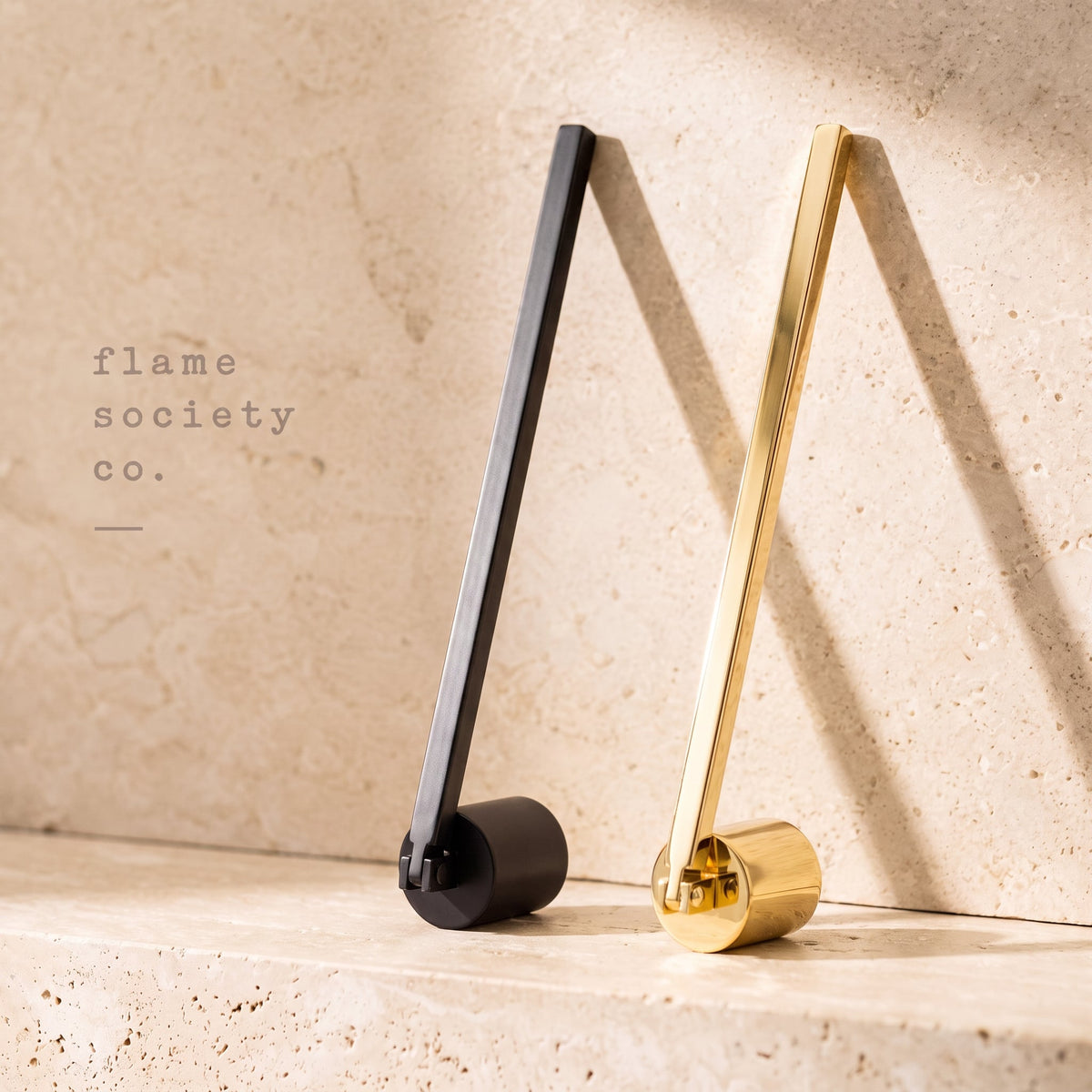 Flame Society Co. wick snuffers in matte black and polished gold leaning against light travertine stone