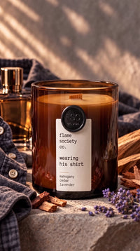 Wearing His Shirt 10oz amber jar candle with wooden wick, styled with a gray shirt, cologne bottle, cedar wood and lavender on stone — mahogany, cedar and lavender — Flame Society Co.