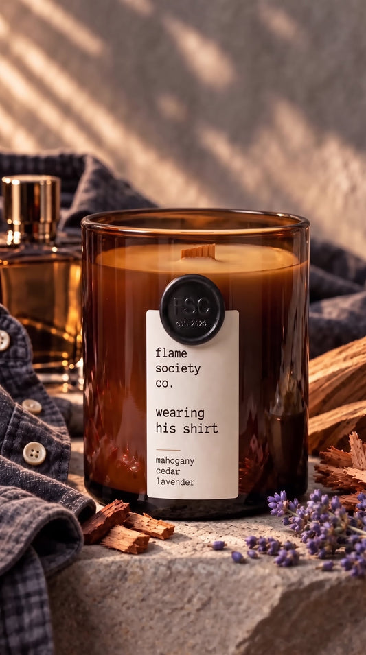 Wearing His Shirt 10oz amber jar candle with wooden wick, styled with a gray shirt, cologne bottle, cedar wood and lavender on stone — mahogany, cedar and lavender — Flame Society Co.