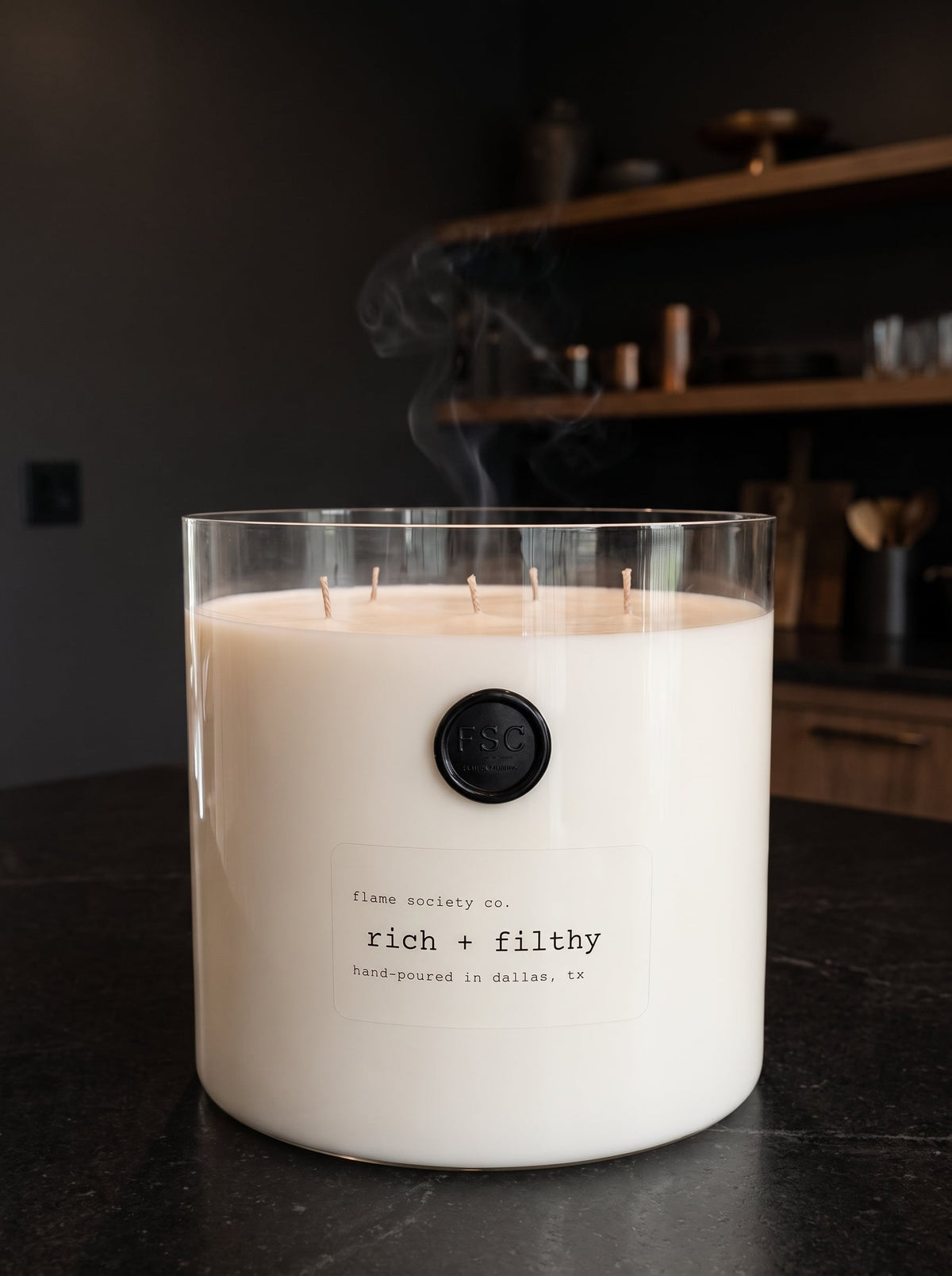 Flame Society Co. oversized rich + filthy statement candle in a clear glass vessel on a black stone island in a moody dark kitchen.