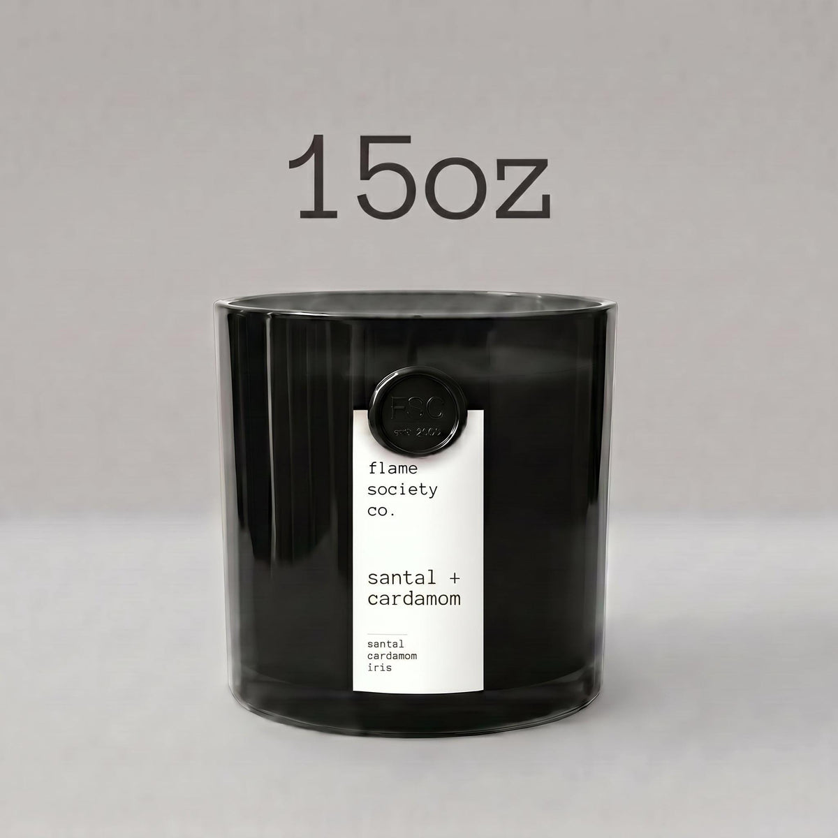 May include: A black cylindrical candle in a glass container with a white vertical label reading Flame Society Co. Santal. The candle is resting on a piece of textured, light brown driftwood against a solid light tan background.