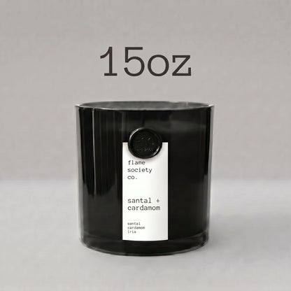 May include: A black cylindrical candle in a glass container with a white vertical label reading Flame Society Co. Santal. The candle is resting on a piece of textured, light brown driftwood against a solid light tan background.