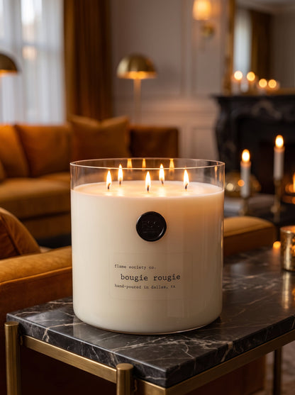 Flame Society Co. Big + Bougie 150oz Bougie Rougie statement candle in a clear glass vessel on a dark marble table in a warm amber living room.
