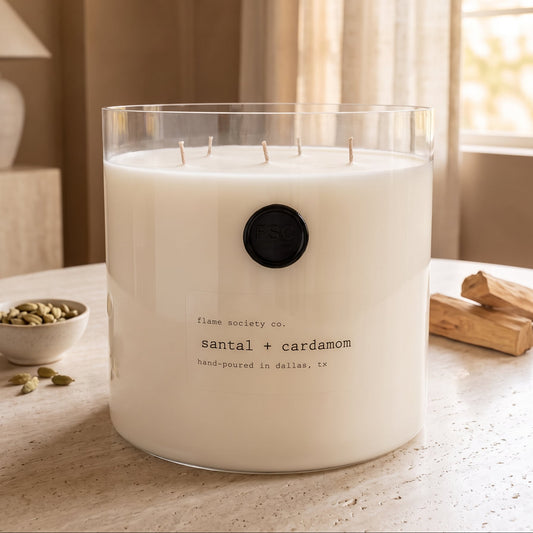 Flame Society Co. Big + Bougie 150oz Santal + Cardamom statement candle in a clear glass vessel on a travertine table with cardamom pods and sandalwood.