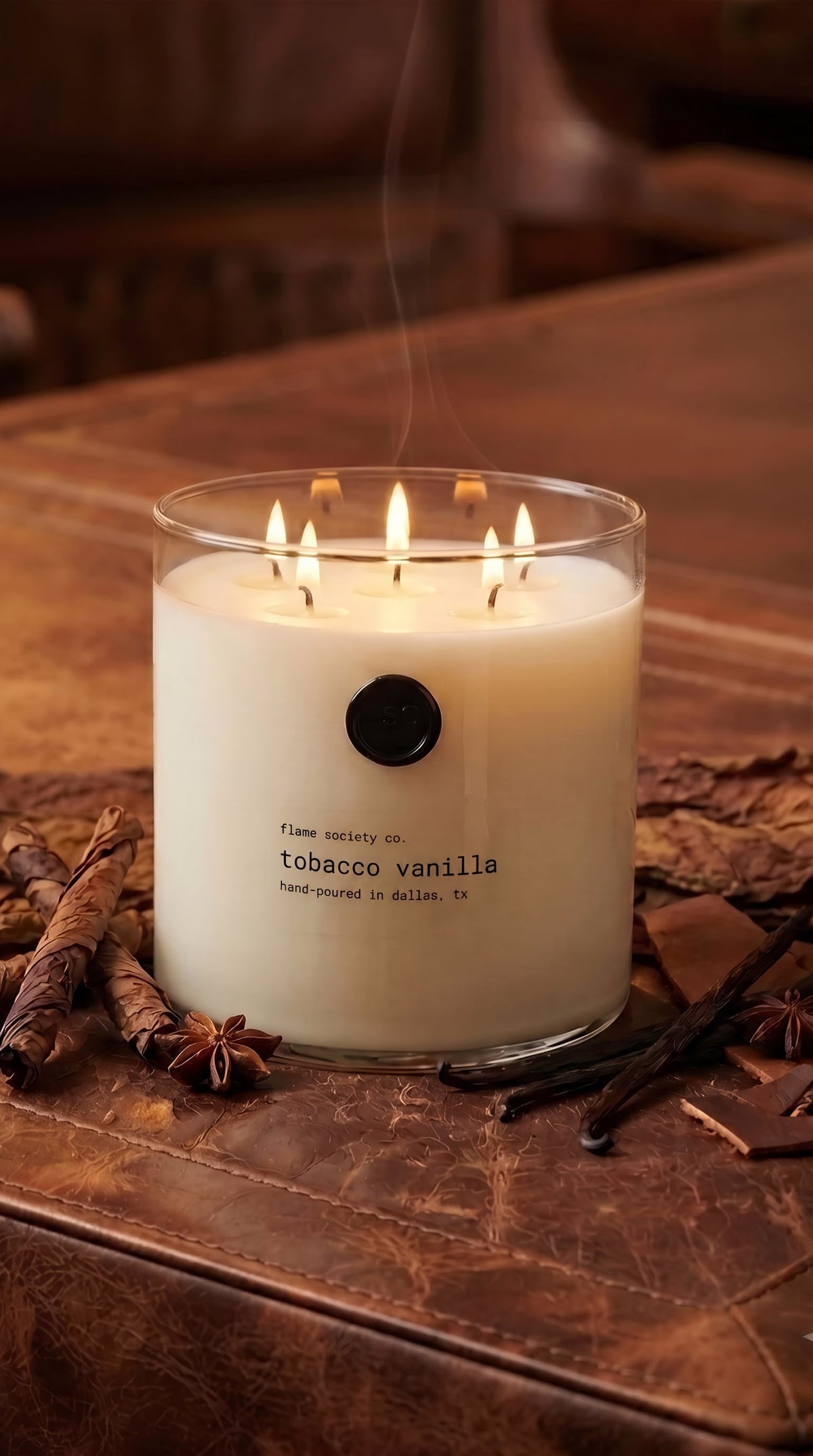 Tobacco Vanilla XL 150oz statement candle in a clear glass vessel on a leather table with tobacco leaves and vanilla — Flame Society Co.