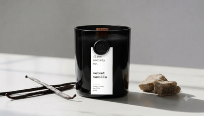 A black glass jar candle with a white label reading 'flavour society co. velvet vanilla' sits on a white surface with vanilla beans and small stones beside it in a minimalist indoor setting.