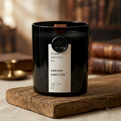 A black glass candle jar with a white label reading 'flame society co. velvet vanilla' and a small wooden wick, styled on a rustic wooden block with blurred books in the background.